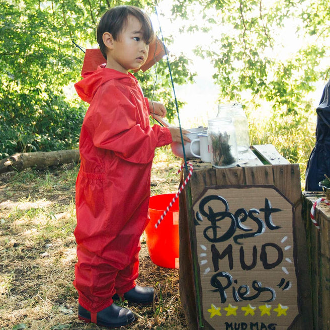 A young child wearing a red waterproof suit, standing at a mud pie stand on a sunny day, looking off to the side.